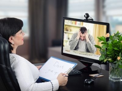Depressed man is exceedingly despondent at first encounter with virtual psychotherapist.  The man sits with his head in his hands. Black-haired glasses psychotherapist watches and hears him on monitor. She holds written message– Dont give up. Horizontal shot indoors