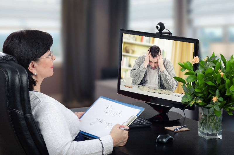 Depressed man is exceedingly despondent at first encounter with virtual psychotherapist.  The man sits with his head in his hands. Black-haired glasses psychotherapist watches and hears him on monitor. She holds written message– Dont give up. Horizontal shot indoors